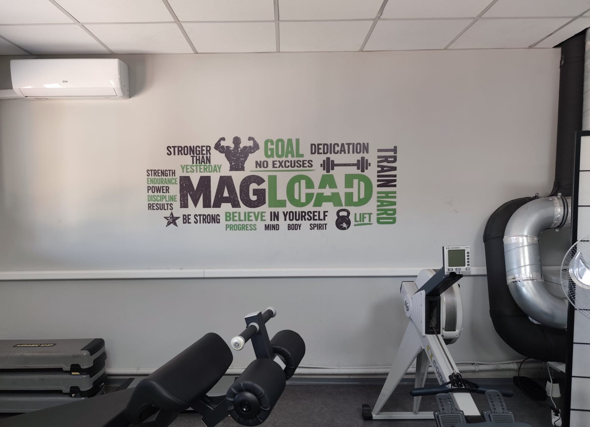 Edzőterem falnyomtatás motivációs szöveggel: MAGLOAD, Stronger, Goal, feliratok és súlyzógép részletei látszanak.