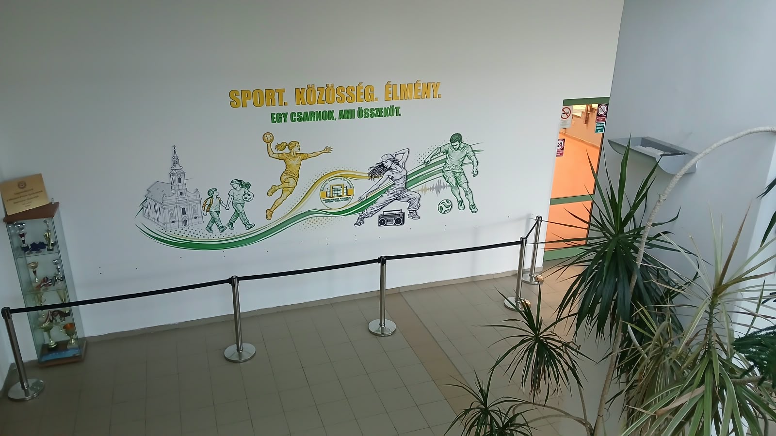 Sport, közösség, élmény feliratú falfestmény futballistákkal, futókkal és templomtoronnyal, beltéri folyosón.