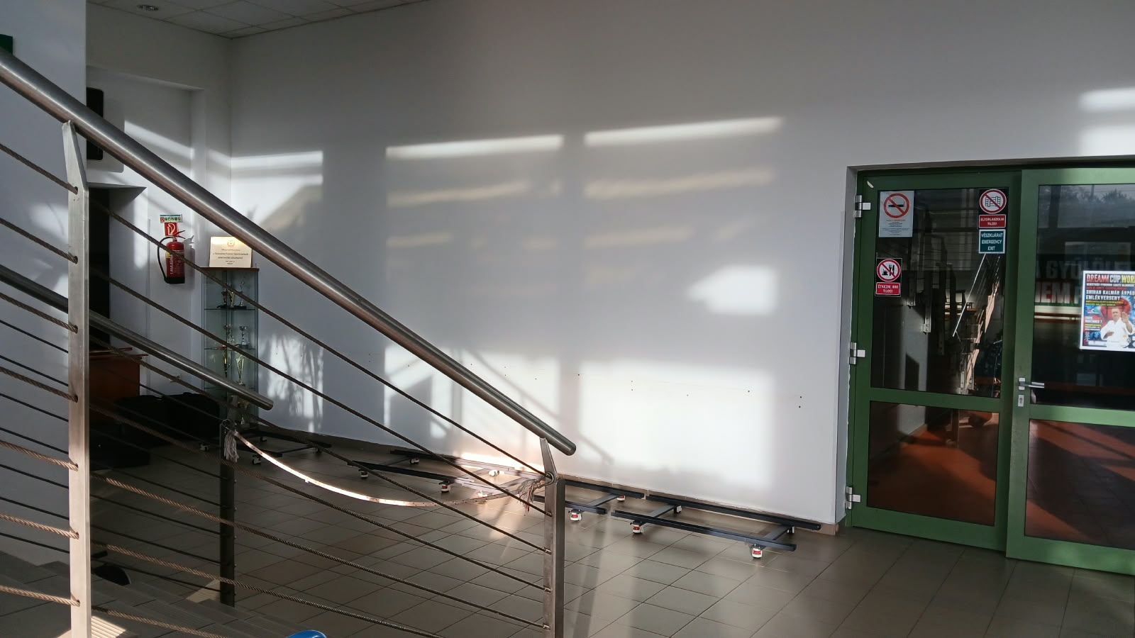 Fehér folyosó, rozsdamentes korlát, rácsos ajtó és zöld keretes bejárat, biztonsági táblákkal.